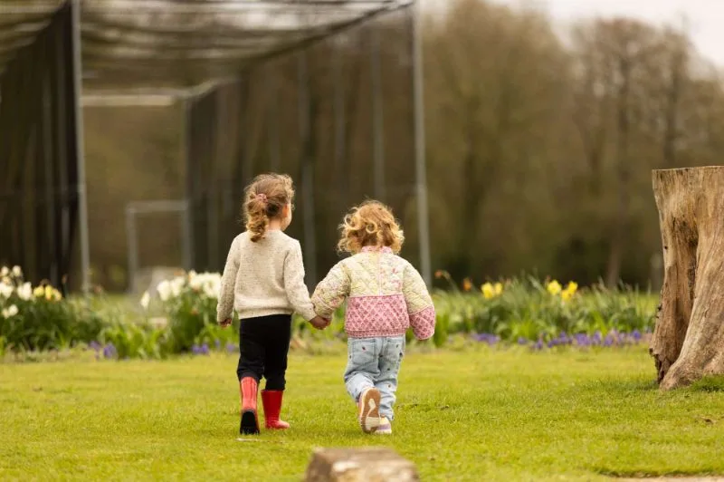 Two young children walking hand in hand across the nursery grounds in wellies